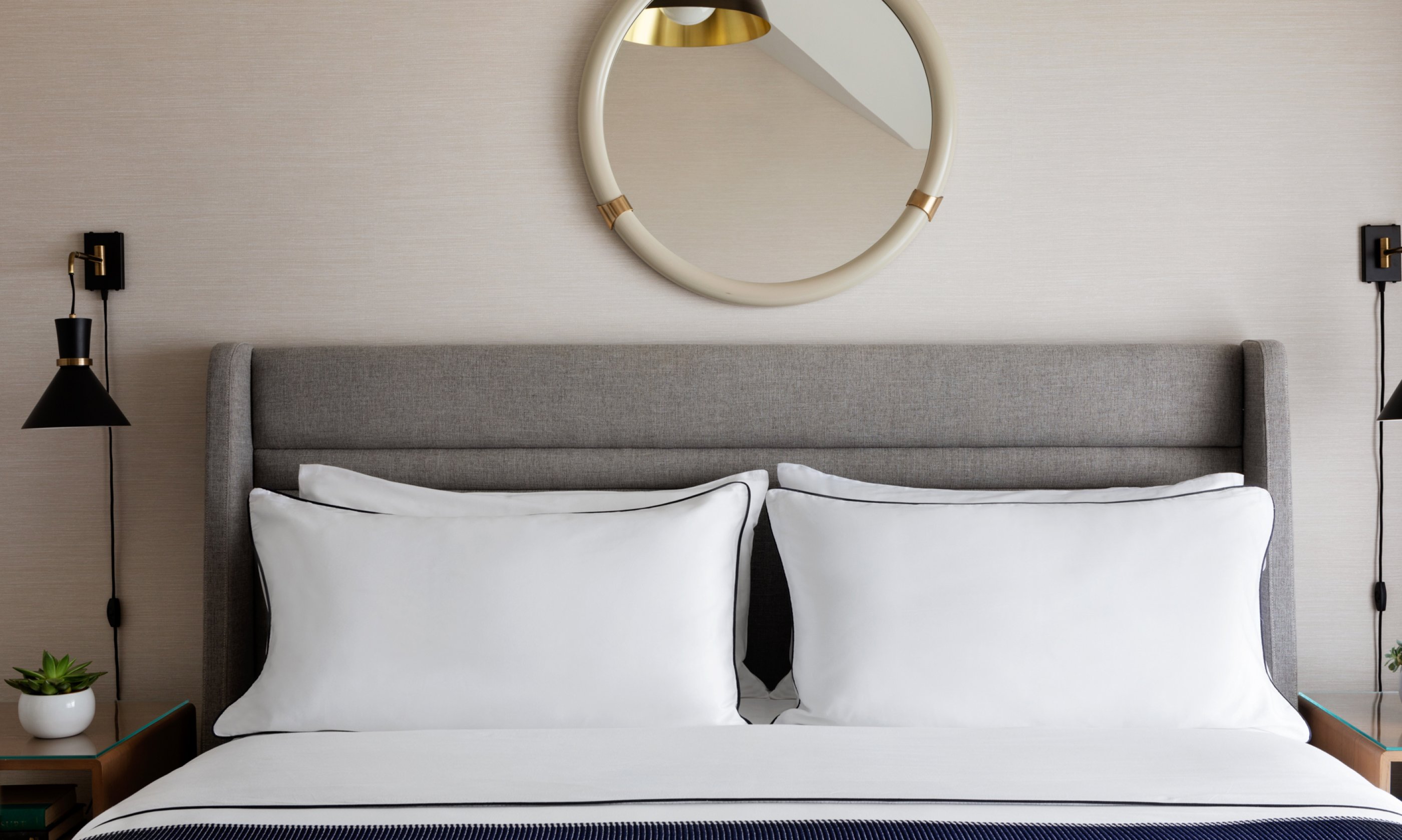 Bed with gray headboard, two large white pillows, a round mirror above, black wall lamps on each side, small plants on nightstands.