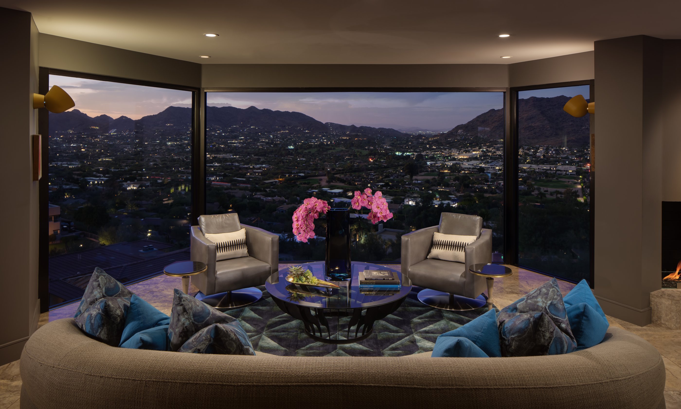 Luxurious living room with large windows showcasing a cityscape view. Two armchairs face a coffee table with pink flowers. Blue pillows on a curved sofa.