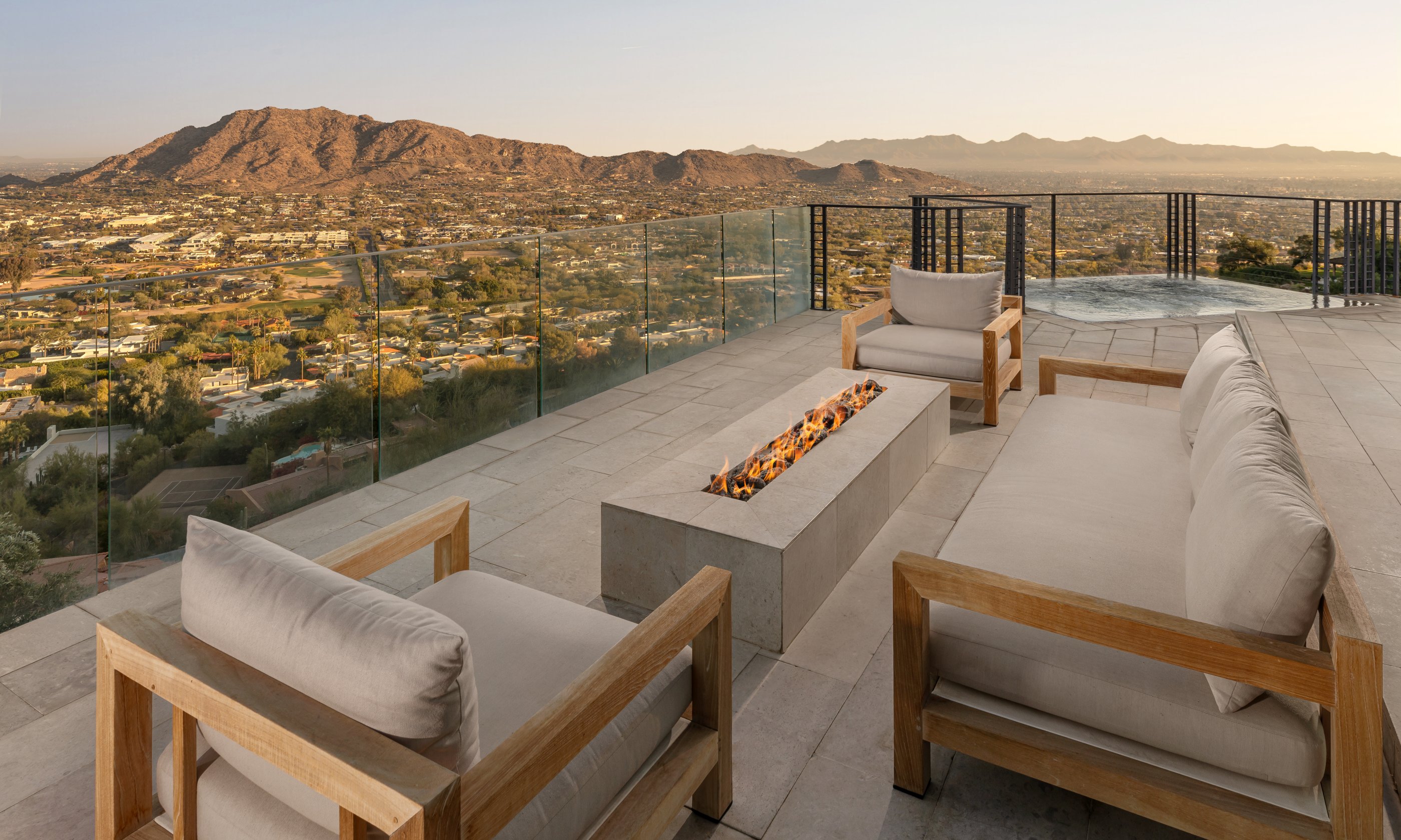 Rooftop patio with wooden lounge chairs and a fire pit, overlooking a cityscape and mountains at sunset.