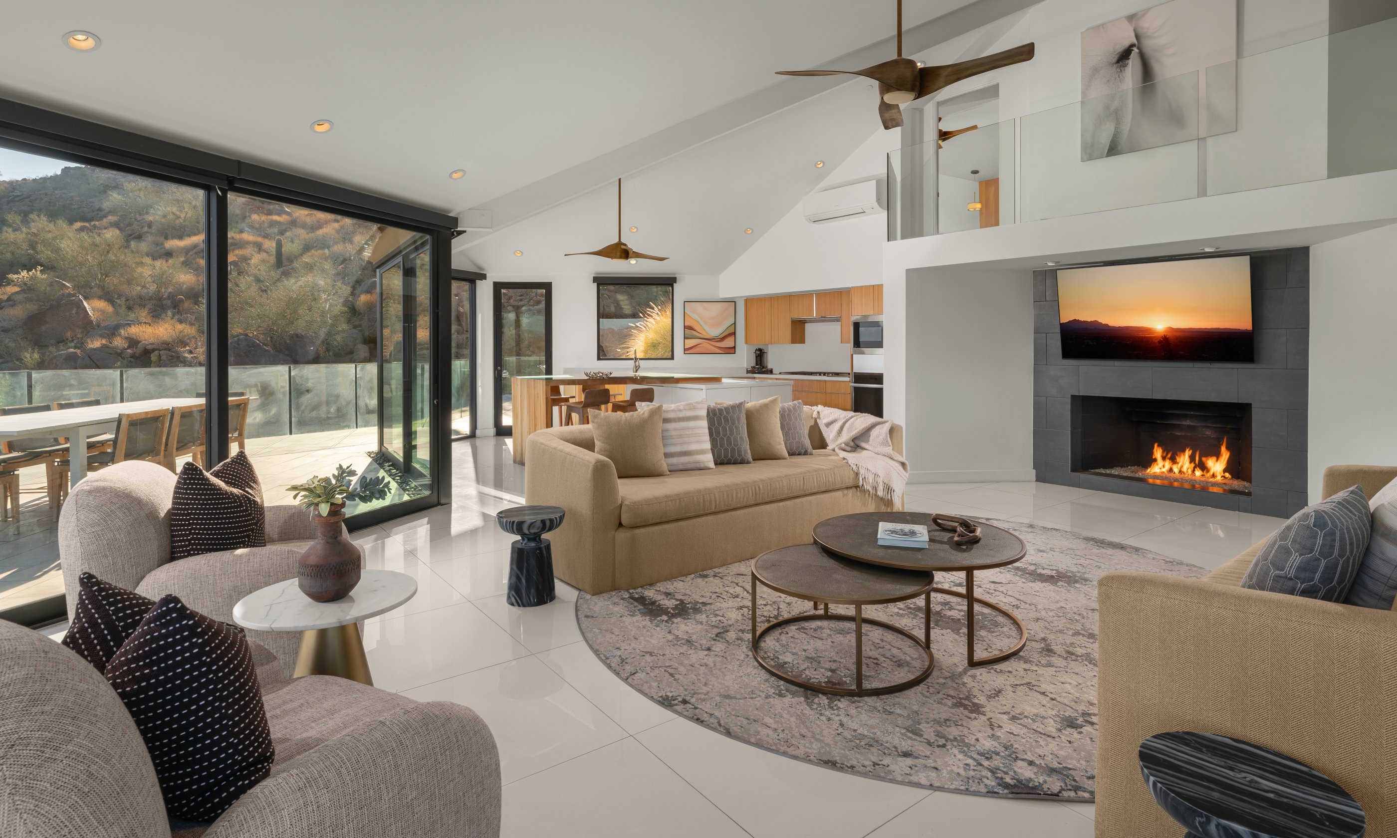 Modern living room with neutral tones, cozy sofas, a round coffee table, large windows opening to a scenic view, and a fireplace with a sunset on TV.
