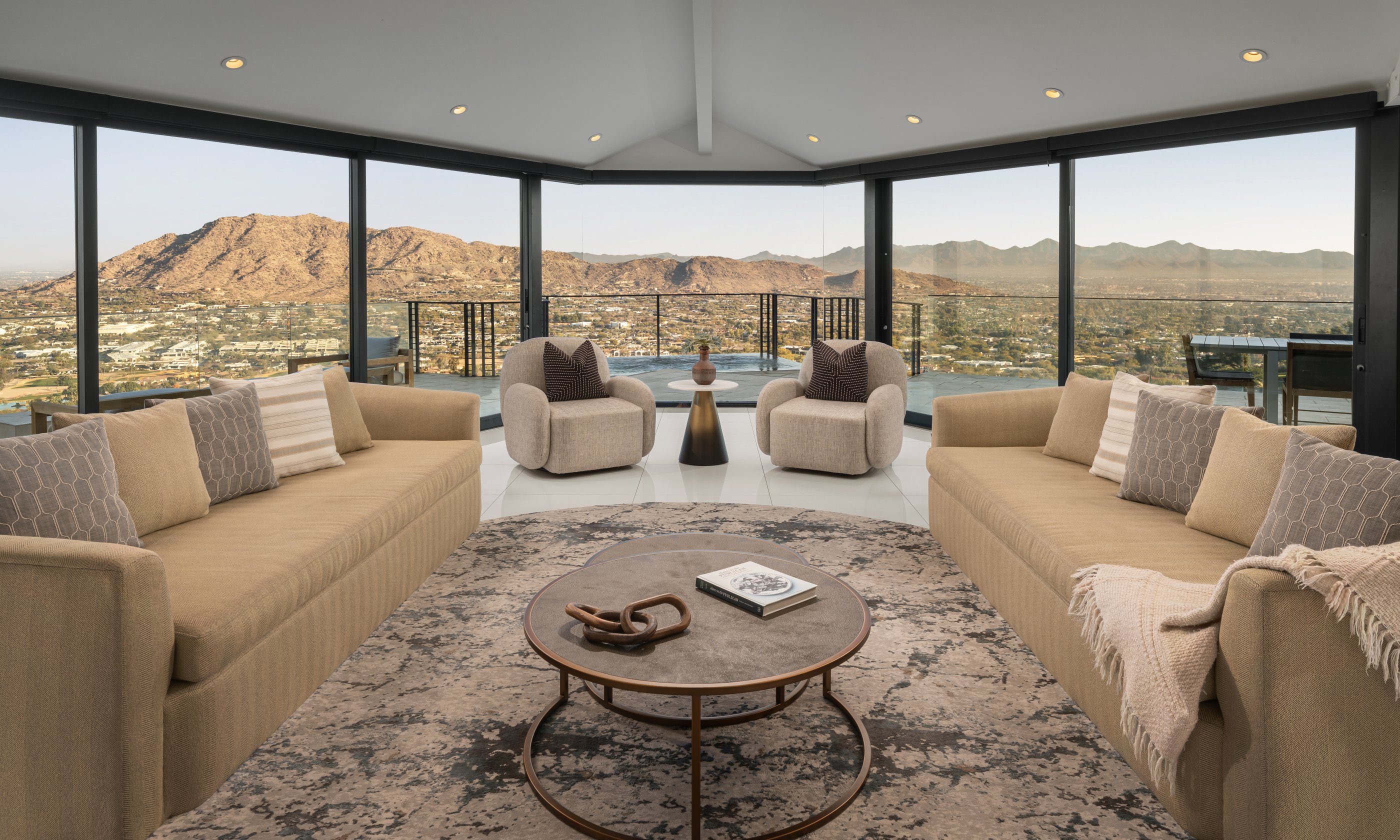 Luxury living room with beige sofas, round table, and large windows offering a mountain view. Neutral tones and cozy ambiance.