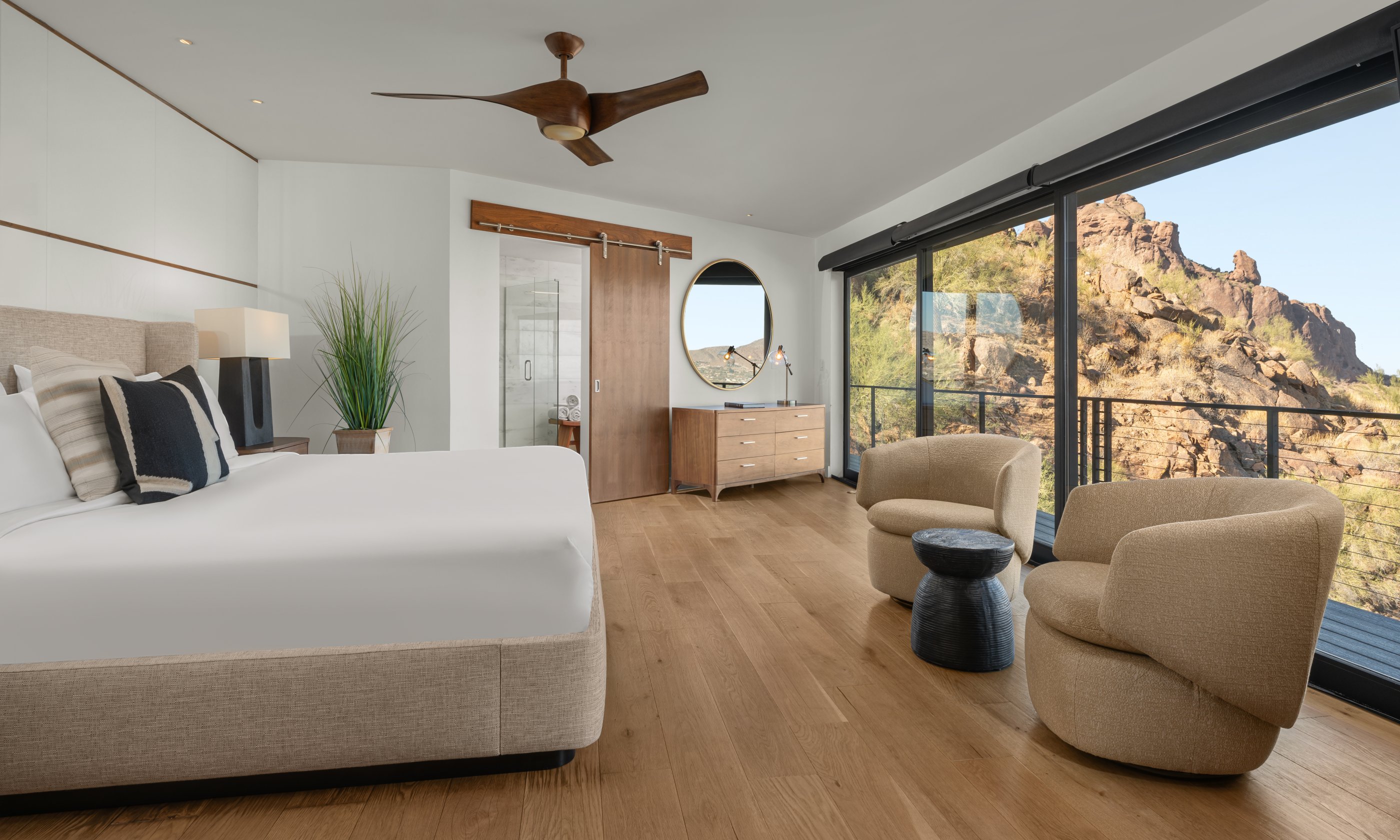 Modern bedroom with a large bed, two beige chairs, and floor-to-ceiling windows offering a scenic mountain view.