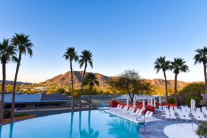 Infinity pool with white lounge chairs and umbrellas surrounded by palm trees with view of sun setting behind the mountain.