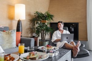 Man in robes sitting by in-room breakfast and using an apple computer.