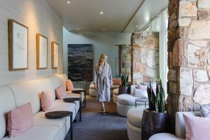 Woman in robe walking through a modern spa lounge with beige sofas, pink cushions, and stone walls.