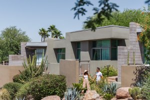 Modern house with geometric design surrounded by desert landscaping, including cacti and rocks. Two people walking in foreground.