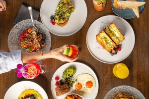 A table with various brunch dishes, including avocado toast, sandwich, and desserts, with a hand reaching for a drink with a lime garnish.