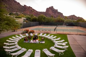 Circular arrangement of lounge chairs on grass, surrounded by desert landscape and mountains at sunset. Tennis courts in the background.