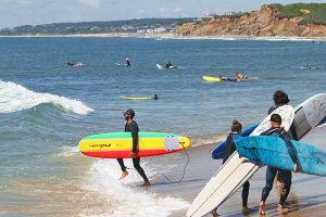 Surfers on a sandy beach carrying colorful surfboards toward the ocean, with waves and rocky cliffs in the background.