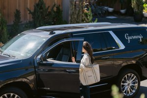 A person with a tote bag is entering a black SUV parked outside a modern wooden house on a sunny day.