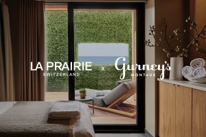 Spa room with a massage table and towels, overlooking a patio with a lounge chair, greenery, and a coastal view. Text: "LA PRAIRIE Gurney’s Montauk."