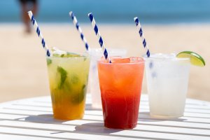 Four colorful cocktails with striped straws on a white table by the beach, with a blurred person walking in the background.