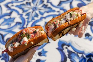Two hands hold lobster rolls over a blue and white patterned table.