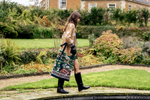 A person walking by a garden pond, wearing a patterned coat and black boots, carrying a large decorative bag. Lush greenery surrounds the area.