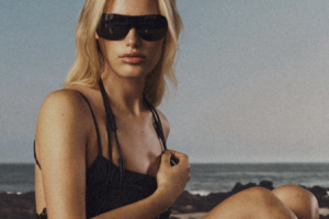 Woman with long blonde hair wearing dark sunglasses and a black outfit, seated on a beach with the ocean in the background.