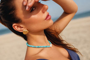 Woman on a beach shielding her eyes from the sun, wearing a blue necklace and gold earrings.