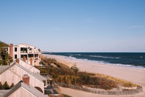 Coastal homes line a sandy beach under a clear blue sky, with gentle ocean waves in the distance.
