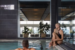 Indoor pool setting with a man swimming and a woman sitting on the edge. Large windows in the background, plants visible. "3 Feet 6 Inch Deep" marked on the pool edge.