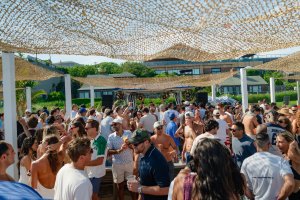 Crowded outdoor party with people mingling under woven canopies. Sunny day with greenery and buildings in the background.