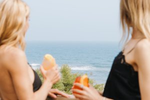 Two women in black dresses enjoy orange drinks on a balcony overlooking the ocean.