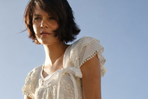 Young woman with short hair in a white dress against a clear blue sky, looking thoughtfully into the distance.