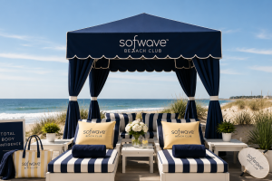 Beach cabana with striped lounge chairs, blue canopy reads "softwave BEACH CLUB," bag and surfboard nearby, ocean and dunes in the background.
