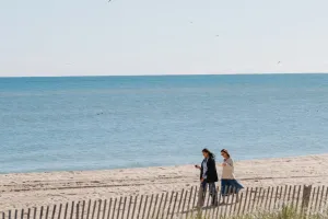 Two people walk along a sandy beach with a wooden fence, while the ocean stretches out under a clear blue sky.