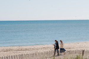 Two people walk along a sandy beach with a wooden fence, while the ocean stretches out under a clear blue sky.