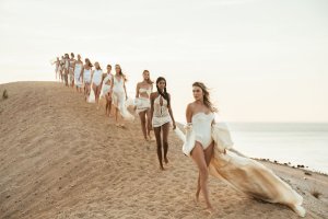 Women in flowing white dresses walking in single file on a sandy dune at sunset.
