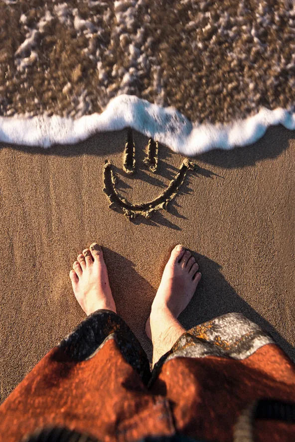Feet on sand near a drawn smiley face, waves approaching.