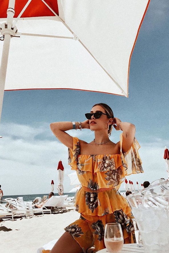 Woman in a floral dress and sunglasses sitting on a beach under a white umbrella, with clear blue sky and ocean in the background.