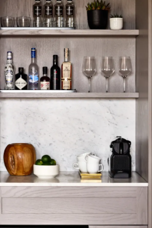 A mini bar with various liquor bottles, glasses, a bowl of limes, a wooden vessel, cups, a coffee machine, and a plant.