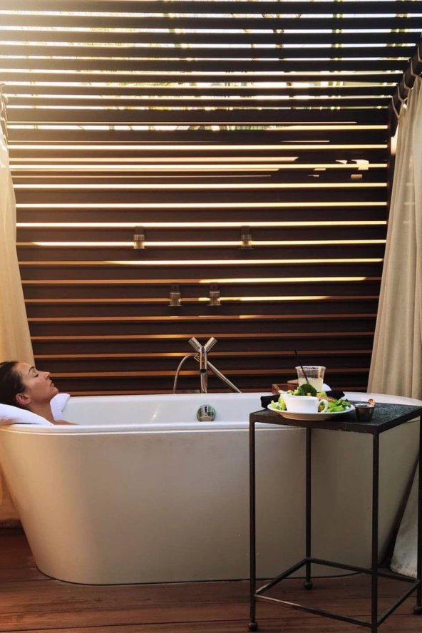 Woman relaxing in an outdoor bathtub under a wooden pergola, with a small side table holding a tea set.