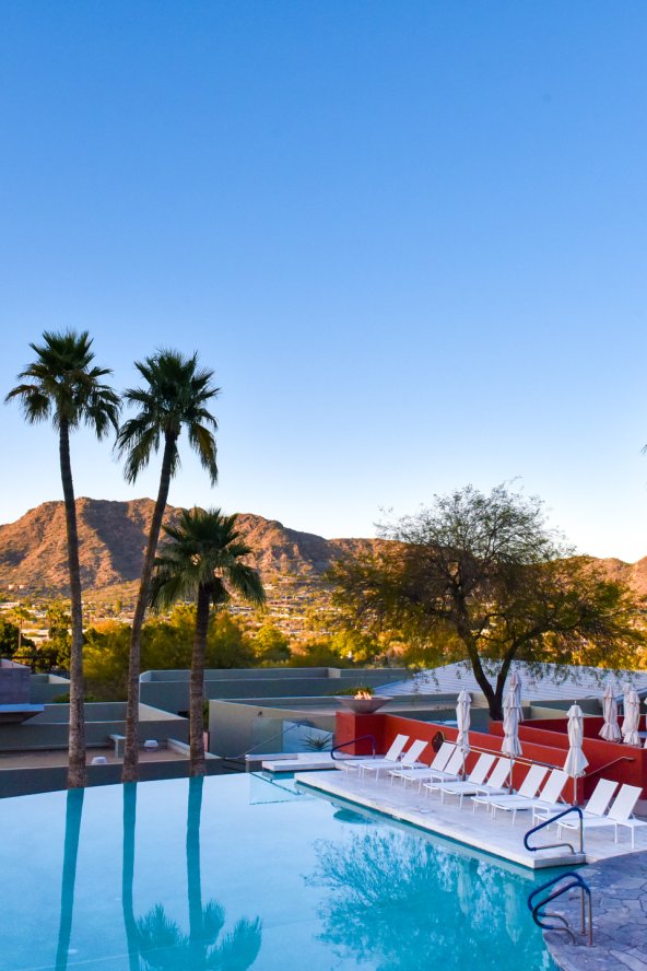 View of Infinity Pool with white lounge chairs and umbrellas as sun sets over mountain in background.