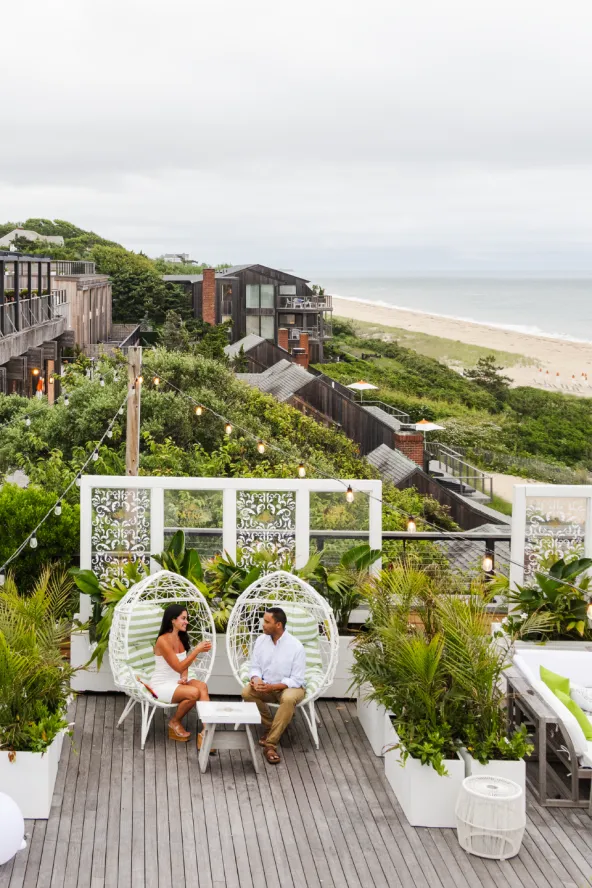 A couple sits drinking cocktails in a plant-filled space with the ocean in the background
