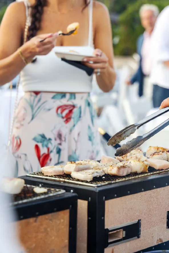 A woman stands in front of a grill as a hand turns clams with tongs