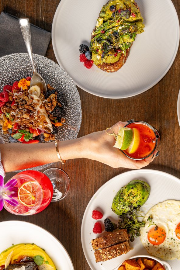 A table with various brunch dishes, including avocado toast, sandwich, and desserts, with a hand reaching for a drink with a lime garnish.