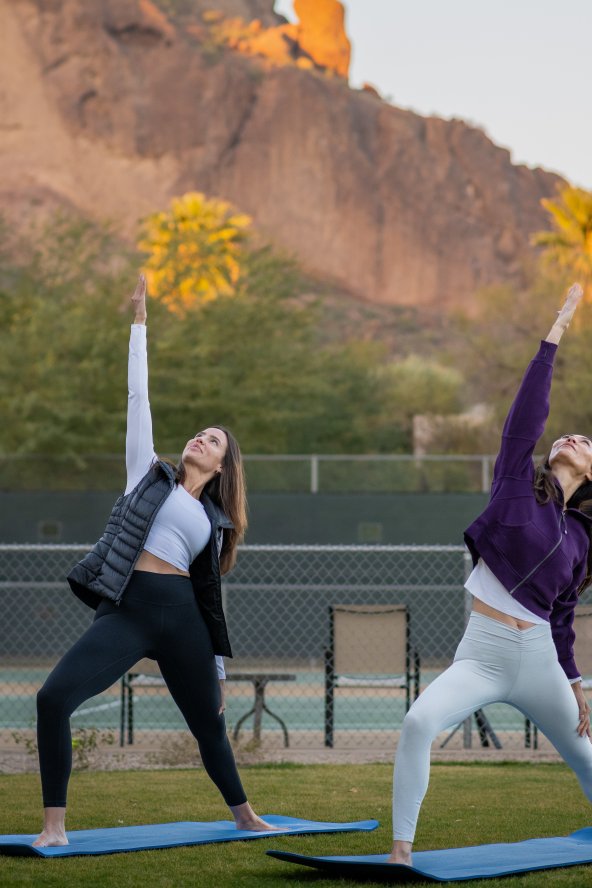 Two women practicing yoga outdoors on mats, performing a side stretch pose. Desert mountains and palm trees in the background.