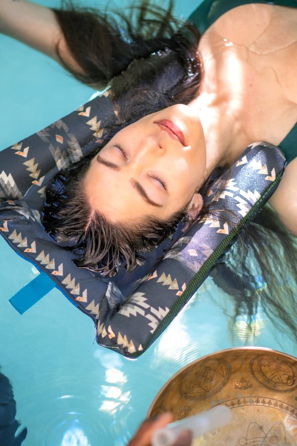 Woman floating in a pool with eyes closed, supported by a cushion, surrounded by two copper bowls.