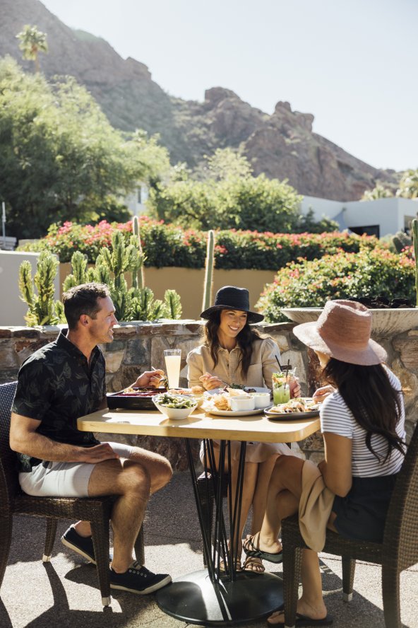 Three people sit at an outdoor patio table enjoying a meal, surrounded by plants and distant mountains under a bright sky.