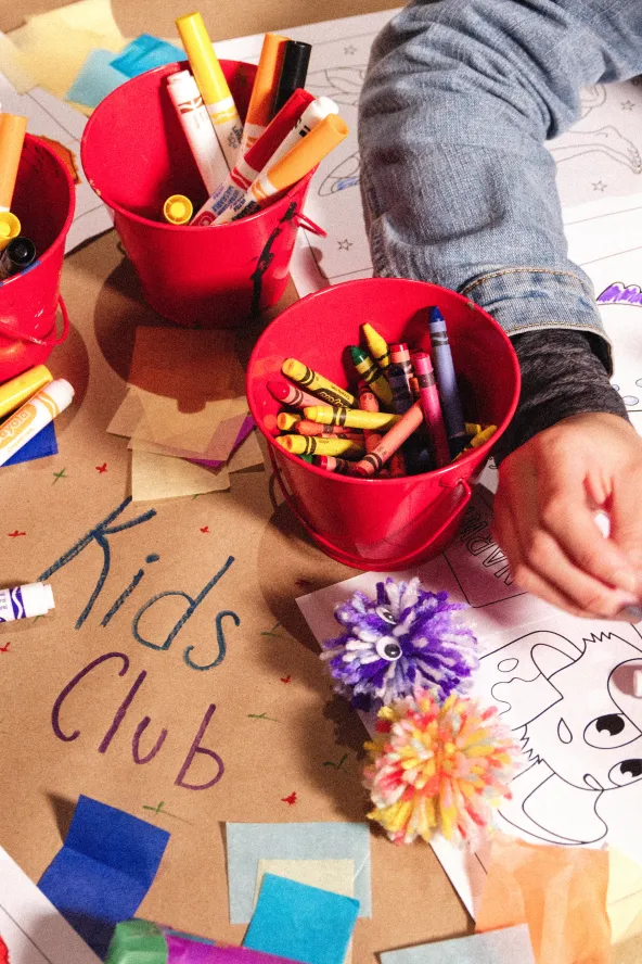 Children coloring at a table with red cups of markers and crayons, surrounded by paper and crafts. Brown paper reads "Kids Club."