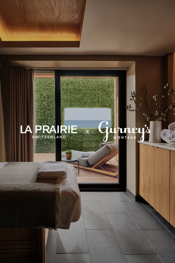 Spa room with a massage table and towels, overlooking a patio with a lounge chair, greenery, and a coastal view. Text: "LA PRAIRIE Gurney’s Montauk."