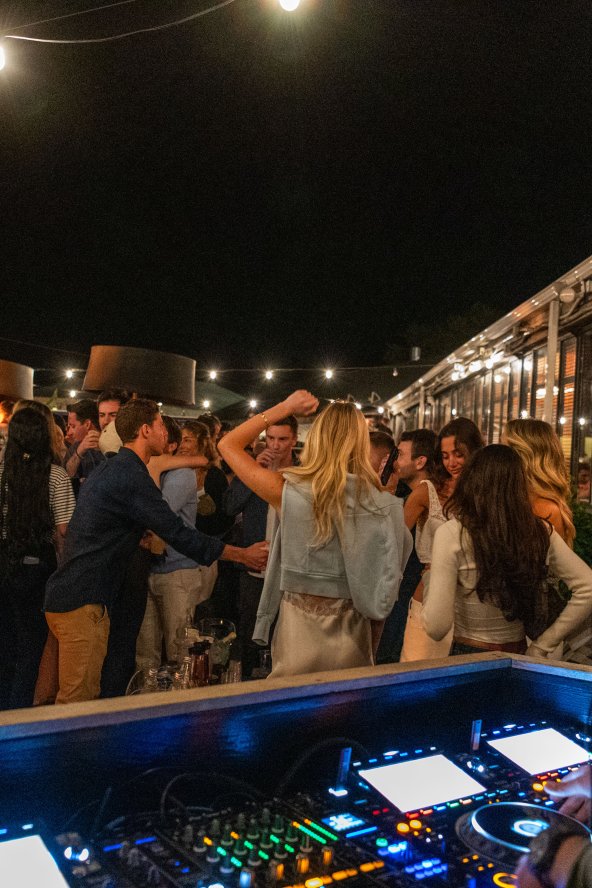 DJ playing music at an outdoor night party, with a crowd dancing under string lights.