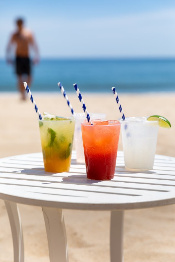 Four colorful cocktails with striped straws on a white table by the beach, with a blurred person walking in the background.