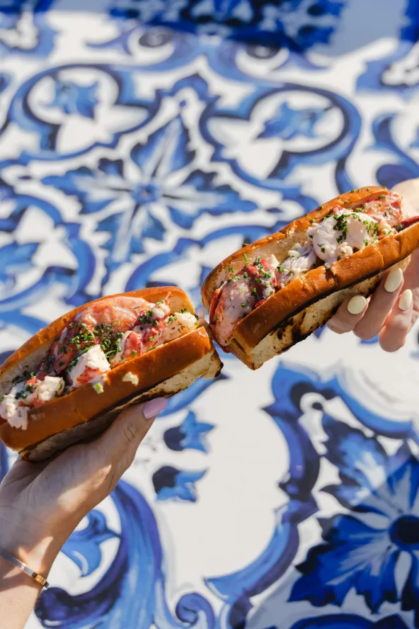 Two hands hold lobster rolls over a blue and white patterned table.