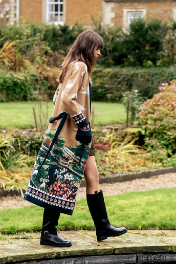 A person walking by a garden pond, wearing a patterned coat and black boots, carrying a large decorative bag. Lush greenery surrounds the area.