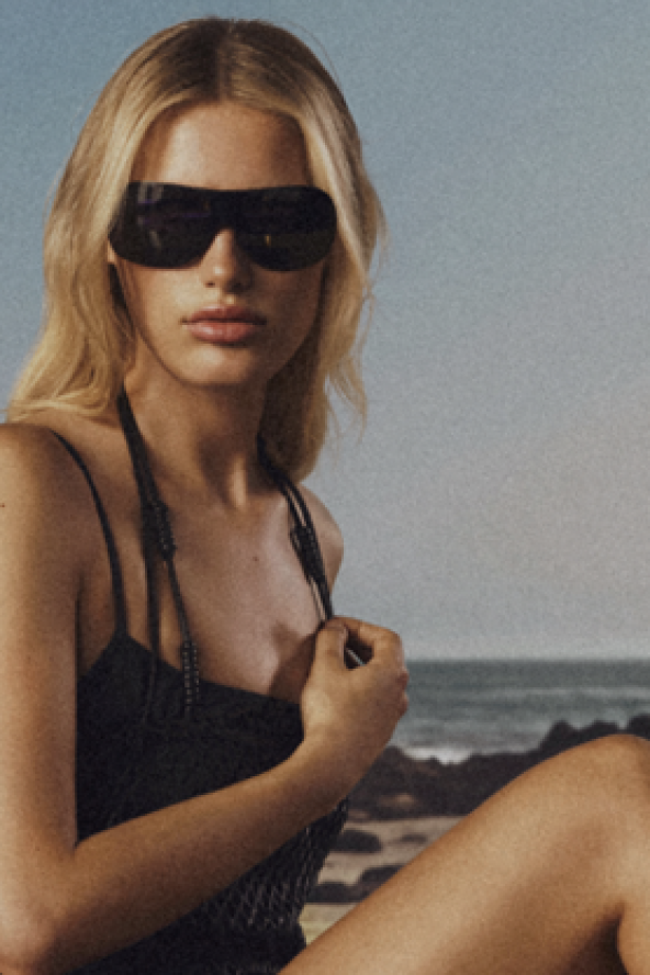 Woman with long blonde hair wearing dark sunglasses and a black outfit, seated on a beach with the ocean in the background.