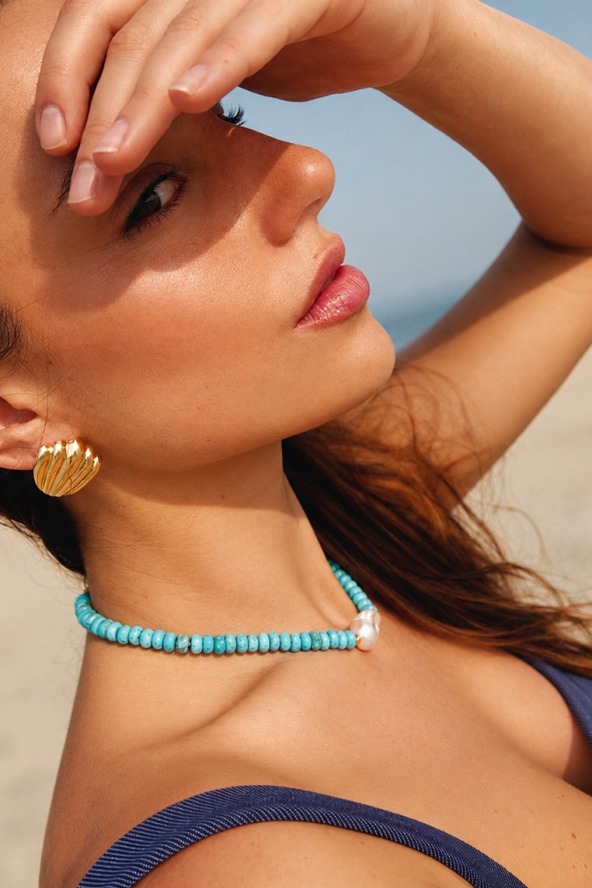 Woman on a beach shielding her eyes from the sun, wearing a blue necklace and gold earrings.