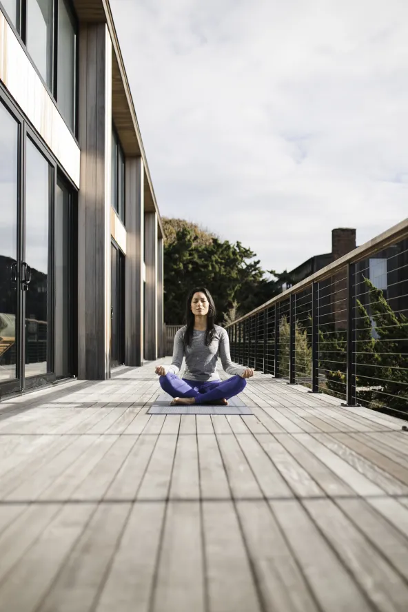 Person meditating on a wooden deck, sitting cross-legged on a yoga mat, surrounded by railings with a building on the left and trees in the background.