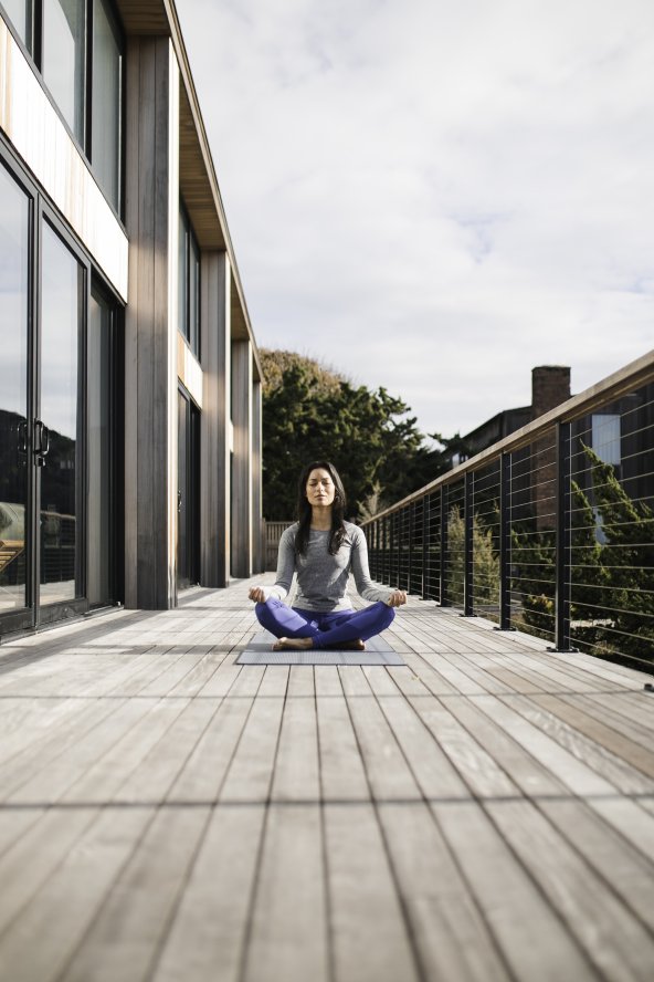 Person meditating on a wooden deck, sitting cross-legged on a yoga mat, surrounded by railings with a building on the left and trees in the background.